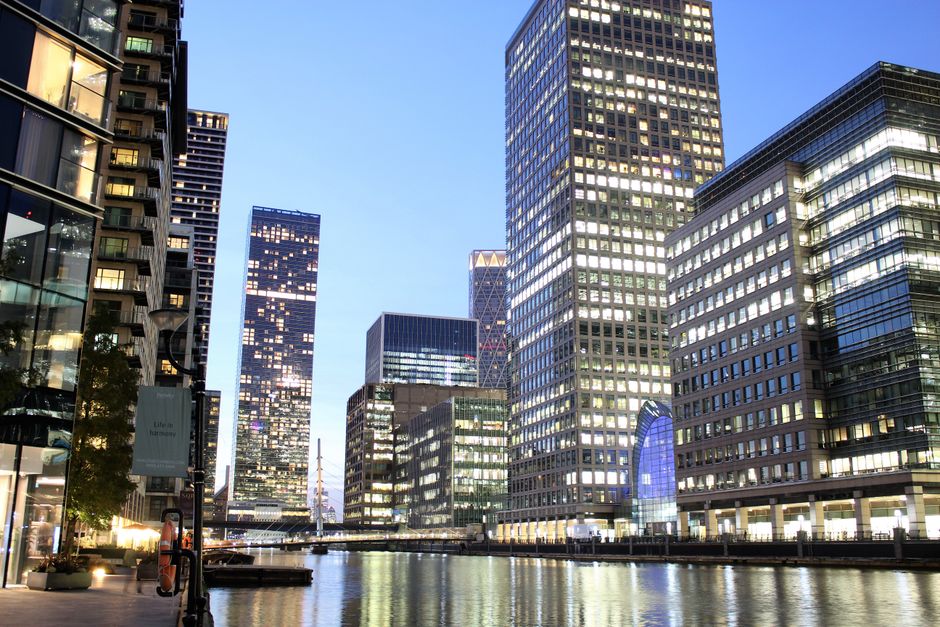 A cityscape featuring modern office buildings with glass facades reflecting the sky, situated along a waterway with a wooden barrier running alongside. In the foreground, a landscaped area includes ne